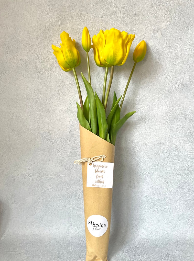 May include: A bouquet of yellow tulips wrapped in brown paper with a tag that reads "happiness blooms from within". The bouquet is tied with twine and has a sticker that says "SDesign Floral".