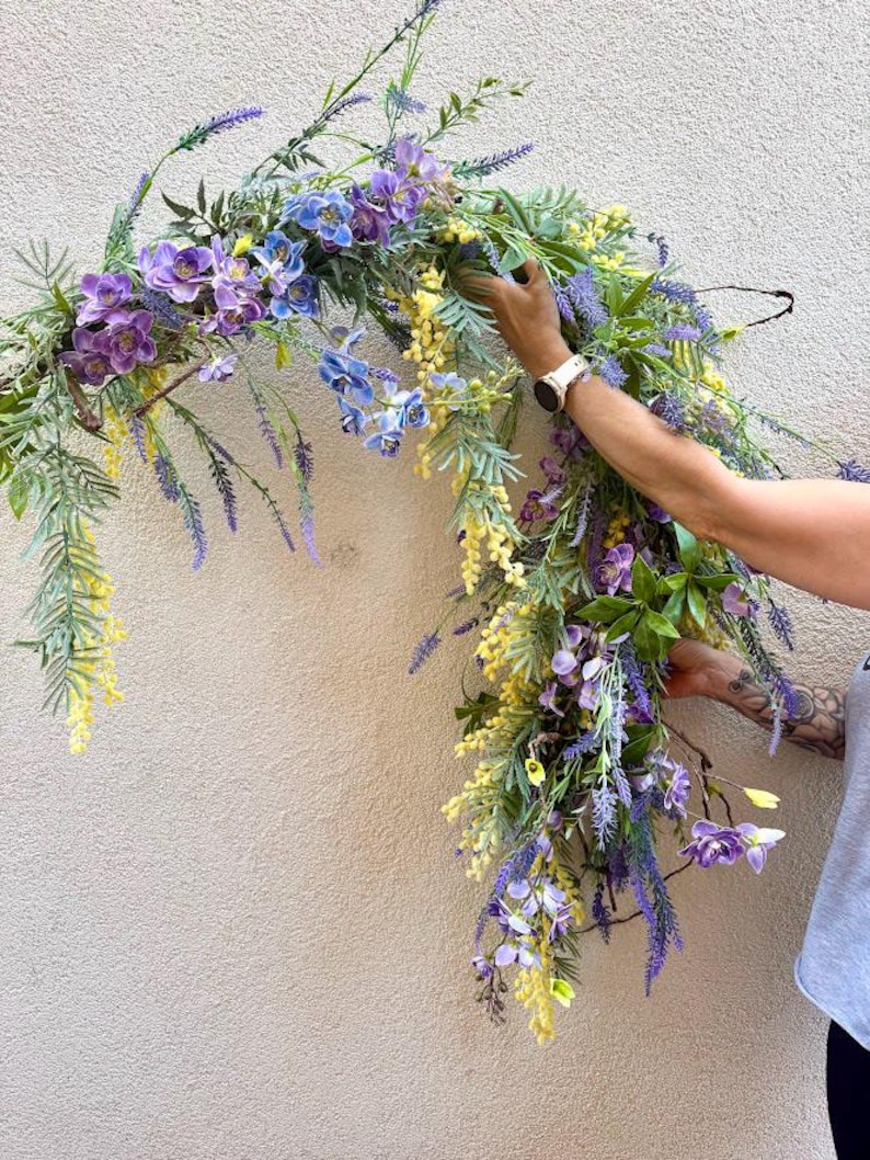 May include: A floral arrangement featuring a variety of artificial flowers in shades of purple, blue, and yellow. The arrangement includes lavender, orchids, and other greenery, creating a natural and vibrant look. The flowers are arranged in a cascading style.