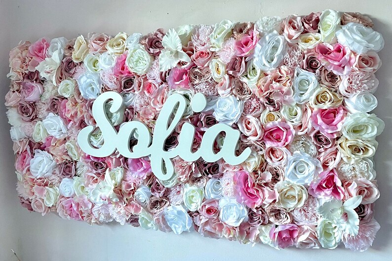May include: A white flower wall with the name "Sofia" spelled out in white letters. The wall is made of a variety of pink, white, and cream flowers.