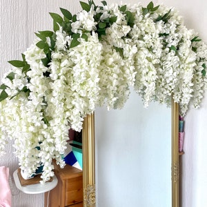 May include: A white artificial wisteria garland draped over a gold framed mirror. The garland is made of white flowers and green leaves.