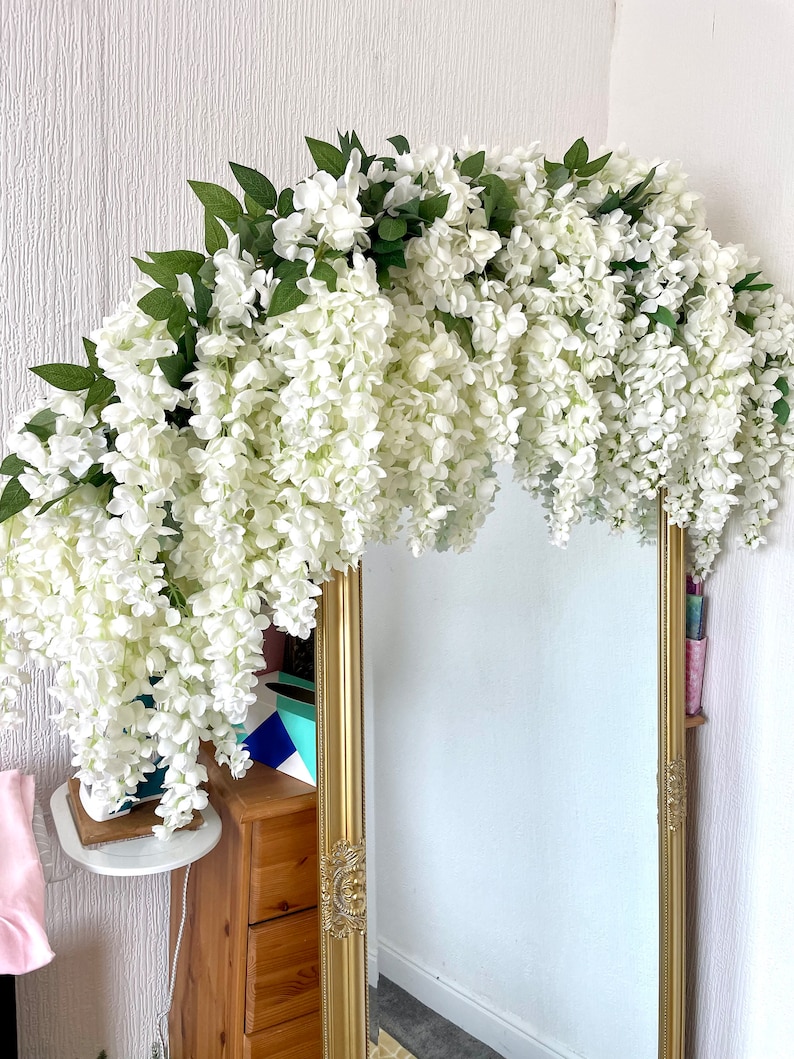 May include: A white artificial wisteria garland draped over a gold framed mirror. The garland is made of white flowers and green leaves.