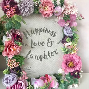 May include: A white picture frame decorated with pink, purple, and green artificial flowers. The frame has the text "Happiness Love & Laughter" printed on it. The picture size is 6" x 8" or 15 x 20 cm.