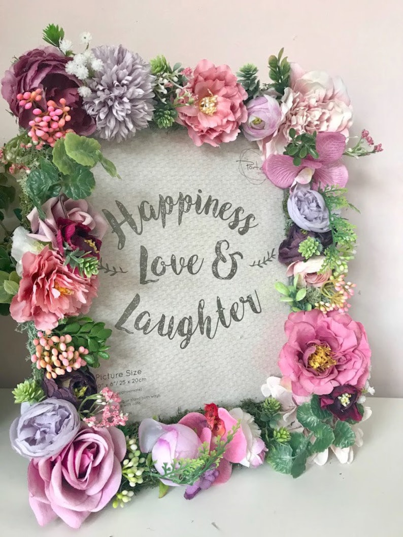 May include: A white picture frame decorated with pink, purple, and green artificial flowers. The frame has the text "Happiness Love & Laughter" printed on it. The picture size is 6" x 8" or 15 x 20 cm.