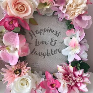 May include: A gray picture frame with a white background and the text "Happiness Love & Laughter" surrounded by a wreath of pink, white, and cream artificial flowers. The picture size is 5" x 7" or 13 cm x 18 cm.