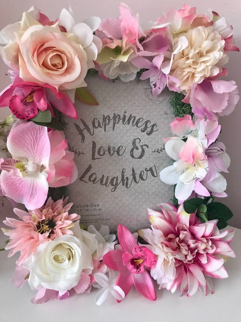 May include: A gray picture frame with a white background and the text "Happiness Love & Laughter" surrounded by a wreath of pink, white, and cream artificial flowers. The picture size is 5" x 7" or 13 cm x 18 cm.