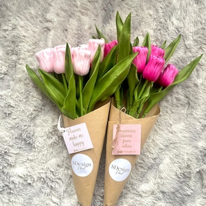 Op de afbeelding: Twee boeketten tulpen verpakt in bruine papieren kegels. E&eacute;n boeket heeft roze tulpen, de andere magenta. Elk boeket heeft een label met de tekst "Flowers make me happy" en "happiness blooms from within", samen met het "SDesign Floral" logo.