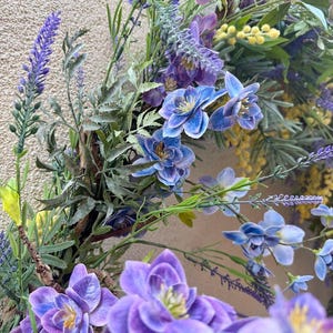 May include: A close-up of a floral wreath featuring a variety of flowers in shades of purple, blue, and yellow. The wreath includes lavender, bluebell-like flowers, and other greenery, creating a natural and vibrant display. The background is a neutral, textured surface.
