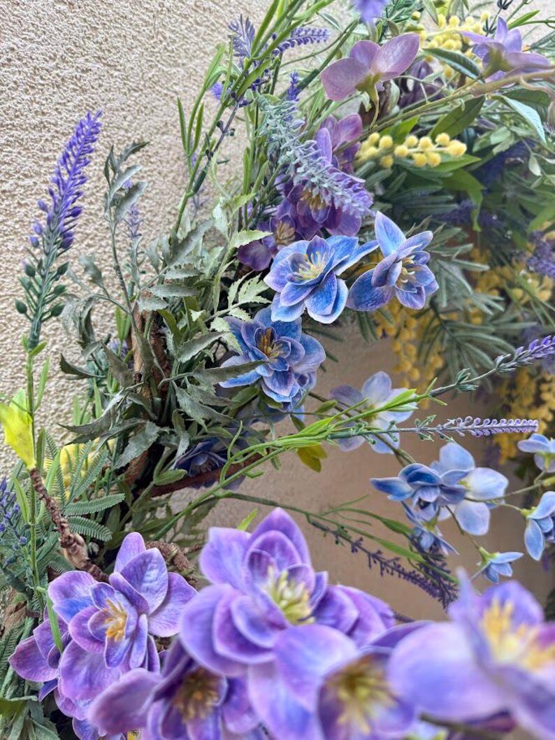 May include: A close-up of a floral wreath featuring a variety of flowers in shades of purple, blue, and yellow. The wreath includes lavender, bluebell-like flowers, and other greenery, creating a natural and vibrant display. The background is a neutral, textured surface.
