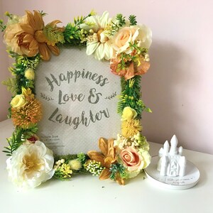 May include: A picture frame decorated with faux flowers in shades of yellow, orange, and white. The frame has a white background with the text "Happiness Love & Laughter" printed in gray. The frame is sitting on a white surface next to a small white ceramic castle.