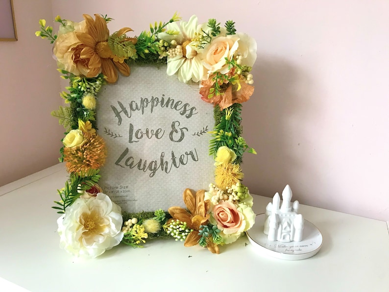 May include: A picture frame decorated with faux flowers in shades of yellow, orange, and white. The frame has a white background with the text "Happiness Love & Laughter" printed in gray. The frame is sitting on a white surface next to a small white ceramic castle.
