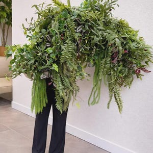 May include: A large, dense floral arrangement featuring a variety of green ferns, leafy branches, and dark berries. The foliage is voluminous and cascading, with long, trailing fronds and deep green leaves, creating a wide, arching botanical display.
