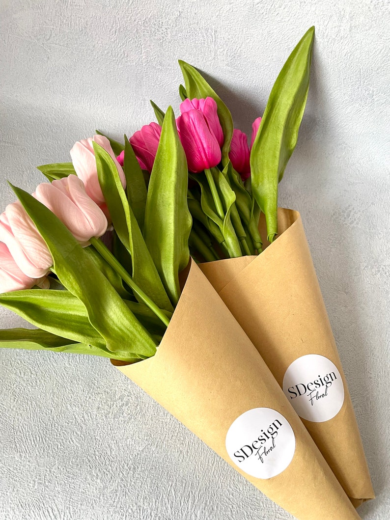 Op de afbeelding: Twee boeketten tulpen, &eacute;&eacute;n met lichtroze bloesems en de andere met levendige fuchsia bloemen. De groene bladeren en stelen zijn verpakt in bruine papieren kegels, elk met een witte sticker met "SDesign Floral".
