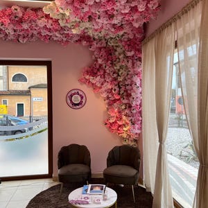 May include: A waiting area with two brown velvet chairs and a white marble table with a gold base. The chairs are facing each other and the table is in between them. The walls are pink and there is a large floral arrangement on the wall behind the chairs. The floral arrangement is made of pink and white flowers. There is a window in the background with a view of a street.