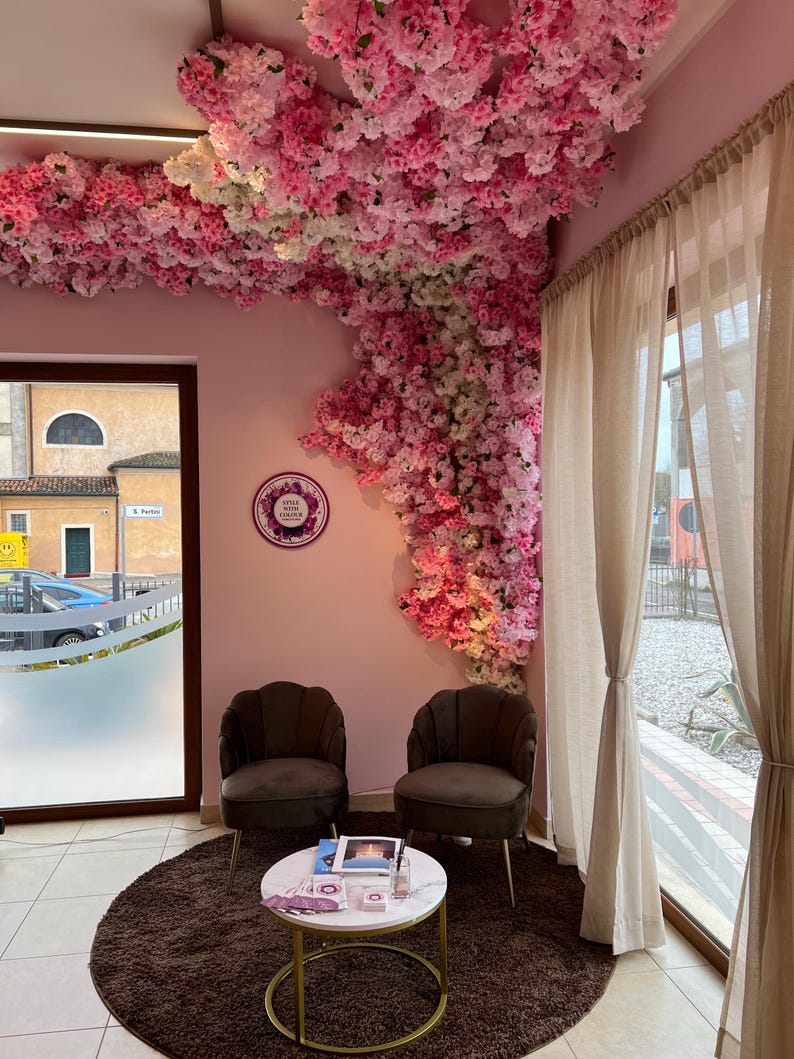 May include: A waiting area with two brown velvet chairs and a white marble table with a gold base. The chairs are facing each other and the table is in between them. The walls are pink and there is a large floral arrangement on the wall behind the chairs. The floral arrangement is made of pink and white flowers. There is a window in the background with a view of a street.