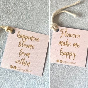 May include: Two pink gift tags with gold lettering. The first tag says "happiness blooms from within" and the second tag says "Flowers make me happy". Both tags have the social media handles "@" and "f" followed by "S Design Floral".