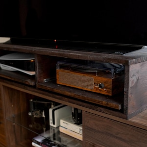 May include: A dark brown wooden entertainment center with a flat screen TV, a record player, and a game console. The entertainment center has two drawers and a glass shelf.