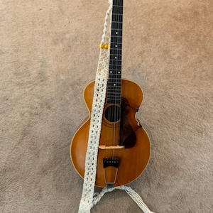 May include: A vintage acoustic guitar with a natural wood finish and a white crocheted strap. The guitar has a soundhole and a pickguard.