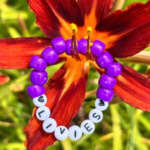 Op de afbeelding: Een paarse kralenarmband met het woord "LIVES" gespeld in witte letterkralen. De armband is versierd met zwarte hartkralen en ligt op een felrode en oranje bloem.