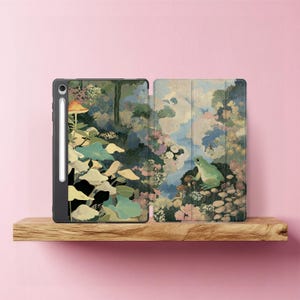May include: A black tablet case with a nature-themed design. The case features a forest scene with a frog, mushrooms, and foliage in shades of green, blue, and pink. A stylus is attached to the side of the tablet.