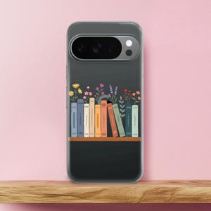 Può includere: Una custodia trasparente per telefono con un design a libreria con libri e fiori colorati. I libri sono di vari colori, tra cui blu, giallo, arancione e verde. I fiori includono fiori gialli, rosa e viola. La custodia è su una mensola di legno su uno sfondo rosa.