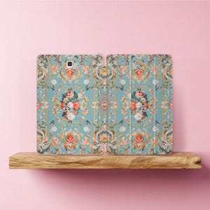 Puede incluir: Una funda para tableta con un diseño floral. La funda está abierta, revelando el interior. El patrón presenta un fondo azul claro con detalles dorados y un ramo de flores en tonos rosa, naranja y blanco. La funda está sobre una estantería de madera.
