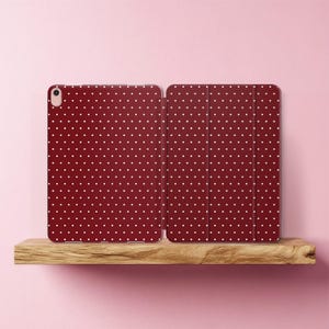 Peut inclure: Une coque de tablette bordeaux avec un motif à pois blancs. L'étui est ouvert, révélant l'intérieur, et est présenté sur une étagère en bois sur fond rose. L'étui a une découpe pour l'appareil photo.