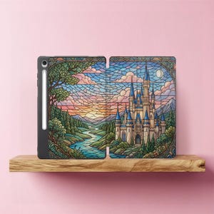 Puede incluir: Una funda negra para tableta con un diseño de vidriera que representa un castillo, un río, árboles y una puesta de sol. La funda está abierta, revelando el interior. Un lápiz óptico blanco está adjunto. La funda está sobre una estantería de madera sobre un fondo rosa.