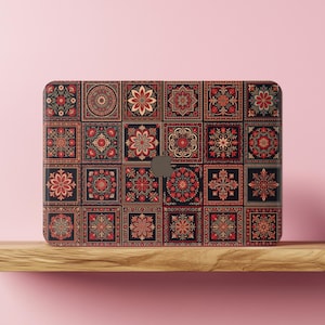 Può includere: Una cover rettangolare per laptop caratterizzata da una griglia di piastrelle quadrate con intricati motivi floreali e geometrici nei toni del rosso, nero e crema. Il design include un ritaglio centrale per un logo circolare.