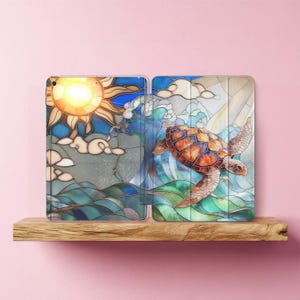 May include: A colorful tablet case featuring a stained-glass design of a sea turtle swimming in the ocean. The case has a sun, clouds, and waves in shades of blue, green, orange, and yellow.