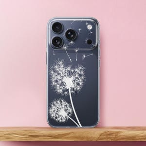 Può includere: Custodia trasparente per telefono con un design di dente di leone bianco su sfondo blu scuro. Il design include due capolini di dente di leone e diversi semi fluttuanti. La custodia è mostrata su una superficie di legno contro uno sfondo rosa.