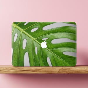 Puede incluir: Una pegatina para portátil con una impresión de alta resolución de una hoja de monstera verde vibrante con sus características perforaciones naturales. El logotipo blanco de Apple es visible en el centro del diseño de la hoja.