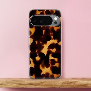 May include: A clear phone case with a tortoiseshell pattern in shades of brown and yellow. The case has a camera cutout and is displayed on a wooden shelf against a pink background.