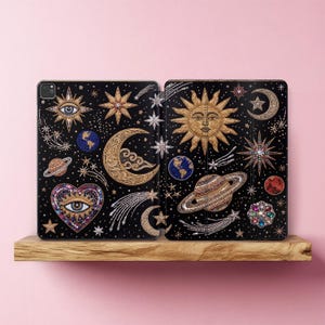 May include: A black tablet case featuring a celestial design with gold, blue, and multicolored beaded embellishments. The pattern includes a sun with a face, crescent moons, planets, stars, comets, and a heart with an eye motif.