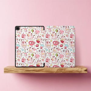 Op de afbeelding: Een tablet hoes en cover met een herhalend patroon van cartoon koeien, aardbeien, melkpakken en drankjes op een witte achtergrond. De hoes is zwart en de cover is wit met hetzelfde patroon. De items worden op een houten plank weergegeven.