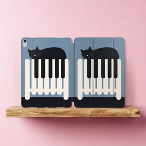 May include: A blue tablet case with a black cat design resting on a piano keyboard. The case is open, showing the interior. The design is a whimsical representation of a cat and piano keys, suitable for music lovers.