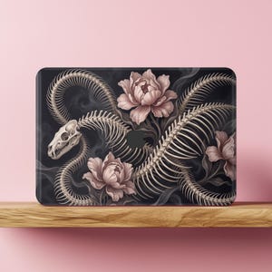 Op de afbeelding: Een rechthoekige laptopskin met een zwarte achtergrond waarop een wit slangenskelet door grote, lichtroze pioenrozen kronkelt. Het ontwerp bevat wazige grijze rookelementen en een centrale uitsparing voor een rond logo-embleem.
