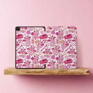 Könnte beinhalten: Eine Tablet-Hülle und -Abdeckung mit einem sich wiederholenden Muster aus rosa Cartoon-Teufeln, Särgen, Pentagrammen und anderen okkulten Symbolen auf weißem Hintergrund. Die Tablet-Hülle ist schwarz.