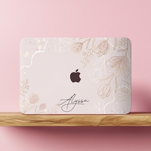 May include: A light pink laptop skin featuring a delicate gold-toned floral line art pattern and the name Alyssa written in a black cursive script. The design includes a central cutout for the Apple logo, surrounded by botanical illustrations.