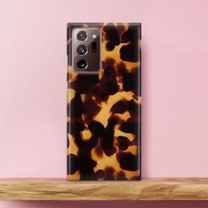 May include: A smartphone case with a tortoiseshell pattern in shades of brown and yellow. The case has a glossy finish and a camera module with three lenses. The phone case is displayed on a wooden shelf against a pink background.