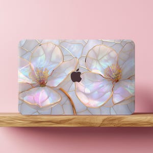 May include: A laptop case featuring a floral design. The case has a white background with a pattern of gold lines. Two large, white flowers with iridescent petals and gold outlines are the focal point. The Apple logo is visible.