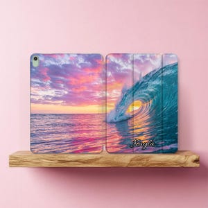 May include: A rectangular tablet case featuring a vibrant sunset over the ocean with a large curling wave. The sky is filled with pink and purple clouds, and the name Kayla is printed in black script on the lower right side of the case.