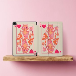Puede incluir: Una funda para tableta con un diseño de carta de la Reina de Corazones. La ilustración muestra a una mujer con pelo rosa, gafas de sol y una flor, sobre un fondo crema con detalles en rosa y naranja. La funda se muestra abierta sobre una estantería de madera.