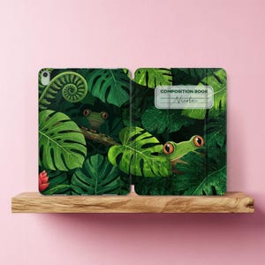 Puede incluir: Una funda para tableta con un patrón de selva verde oscuro con hojas de monstera, helechos y dos ranas de ojos rojos. El lado derecho incluye una etiqueta blanca con el texto COMPOSITION BOOK y el nombre Nicole escrito en letra cursiva.