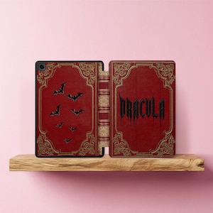 May include: A red tablet case with gold filigree and black bat silhouettes. The right side of the case features the word "DRACULA" in a gothic font. The case is open, revealing the spine of a book.