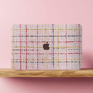 May include: A laptop skin featuring a woven fabric design in shades of pink, red, yellow, and blue. The skin has a grid pattern and a dark brown Apple logo in the center. The laptop skin is on a wooden shelf against a pink background.