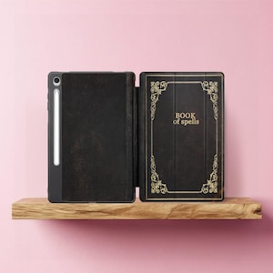 Peut inclure: Étui noir pour iPad avec un design de livre vintage. L'étui est ouvert, révélant l'intérieur et le texte "BOOK of spells" en lettres dorées. L'étui est posé sur une étagère en bois sur fond rose.