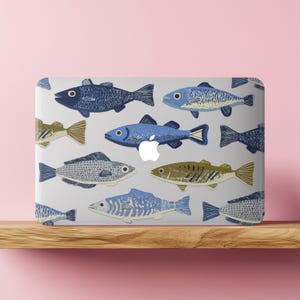 May include: A laptop with a gray surface featuring a pattern of various fish illustrations in shades of blue, green, and tan. The Apple logo is visible. The laptop is resting on a wooden shelf against a pink background.
