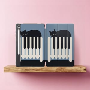 Könnte beinhalten: Eine schwarze Tablet-Hülle mit blauem Hintergrund, die eine schwarze Katze zeigt, die sich auf einem Klaviertasten-Design ausruht. Die Hülle ist geöffnet und zeigt den Tablet-Bildschirm und einen Stift. Das Design wiederholt sich auf dem Cover.