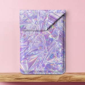 Puede incluir: Una funda para portátil holográfica con un patrón iridiscente morado, azul y blanco. La funda está hecha de un material suave y duradero y está diseñada para proteger tu portátil de arañazos y golpes.