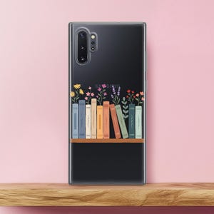 May include: A clear phone case featuring a black background with a bookshelf design. The bookshelf has colorful books and flowers on top. The phone case is on a wooden surface with a pink background.